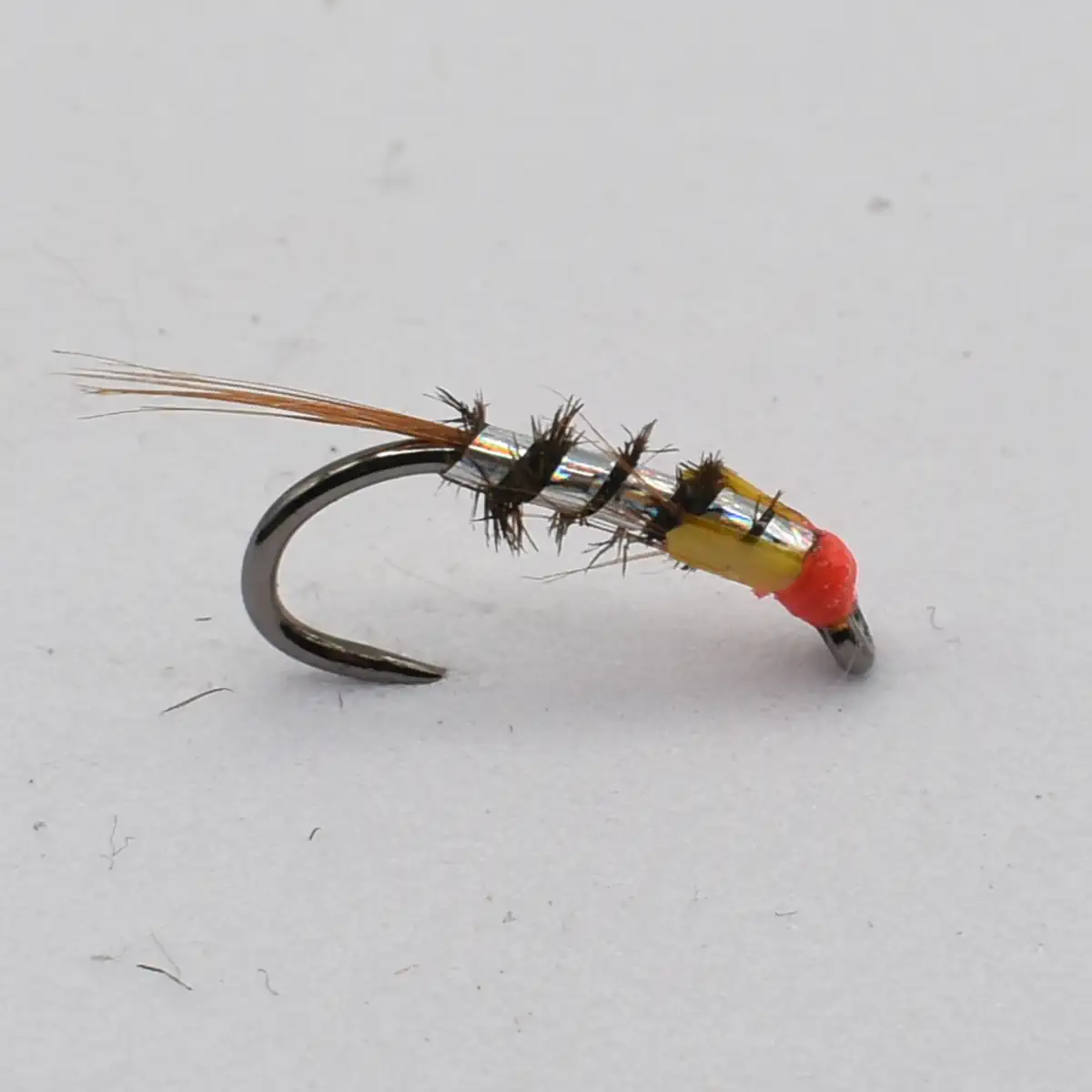 Close-up of a small fishing fly: curved hook with an orange bead head, yellow body wrap, black hackle, and a long tan tail.