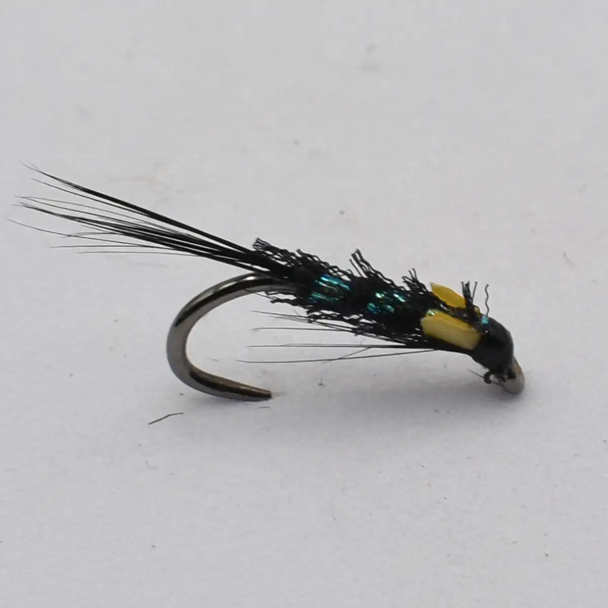 Close-up of a black fishing fly with long hair-like tail fibers, turquoise beads, and a yellow bead near the head on a curved hook.
