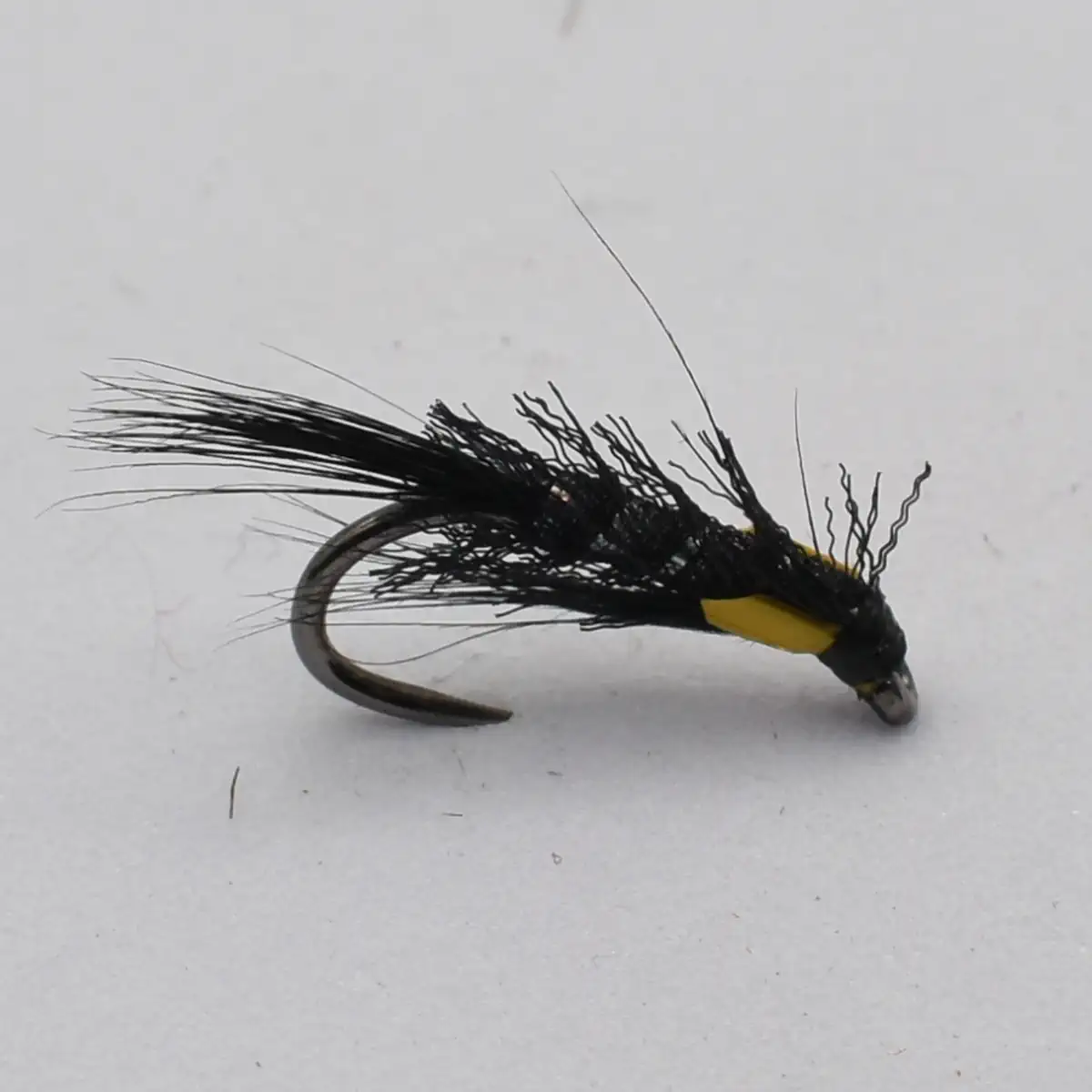 Artificial fishing fly with black hackle and a yellow head mounted on a curved hook against a white background, used as a lure for trout fishing.