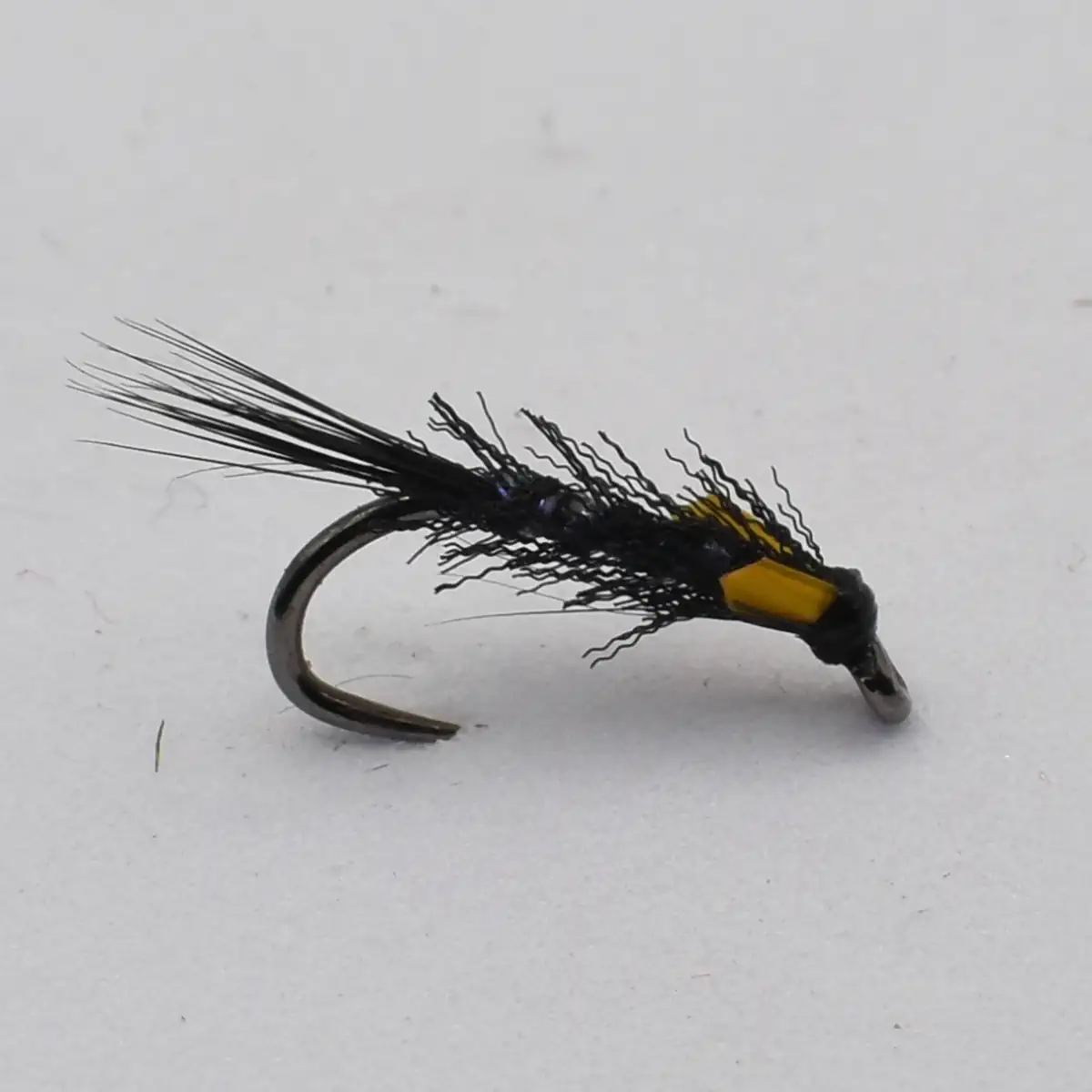 Black fishing fly with wispy black hackle and yellow tag tied to a curved hook.