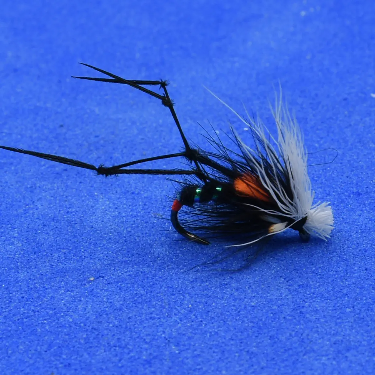 Black fishing fly lure with orange head and white-tipped tail laid on a blue background; used for fly fishing