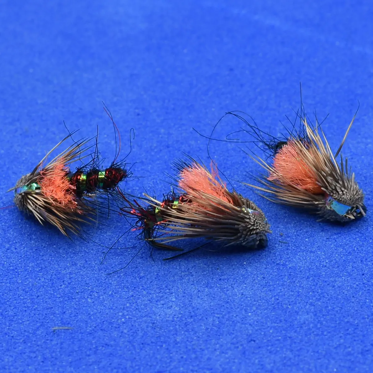 Three hand-tied fishing flies with orange and gray feather bodies on a blue background.