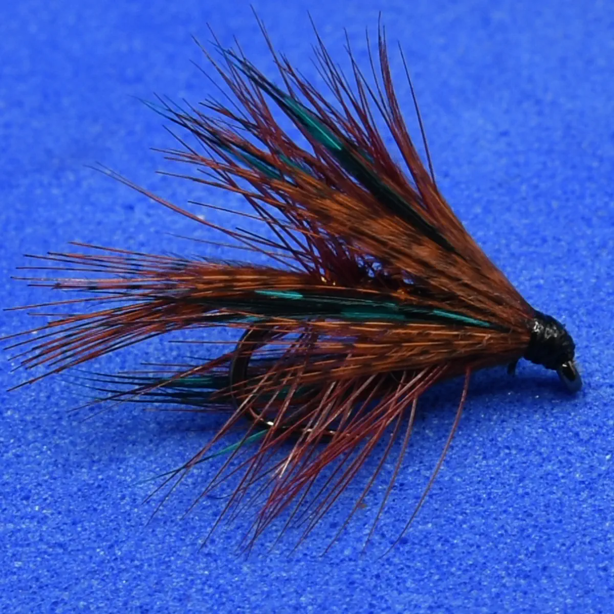 Artificial fishing fly with brown and green hackle on a blue background, used as a lure for fly fishing.