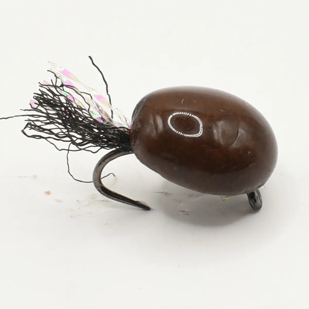 Brown egg-shaped fishing lure with a curved hook and dark fiber tail, shown from the side with a small reflective dot on its surface.