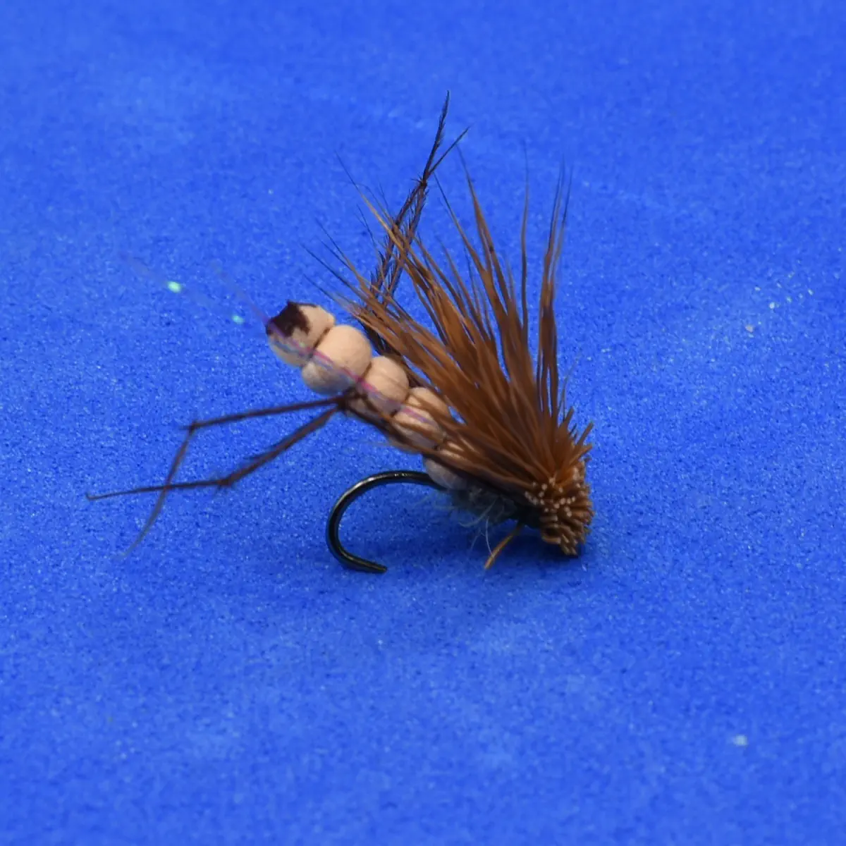 Artificial fishing fly with brown hackle and segmented beige body on a curved hook, lying on a blue background for fly fishing use.