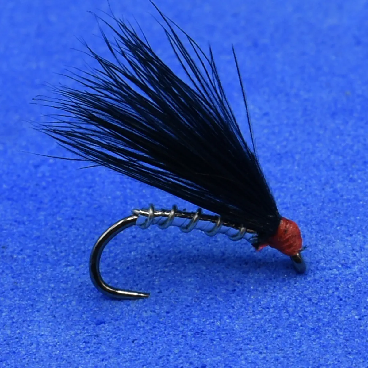 Close-up of a black feathered fishing fly with a red head tied to a curved hook on a blue background.