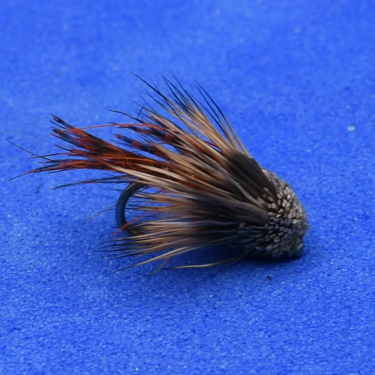 Artificial fly fishing lure with brown and tan feather fibers on a curved hook resting on a blue surface