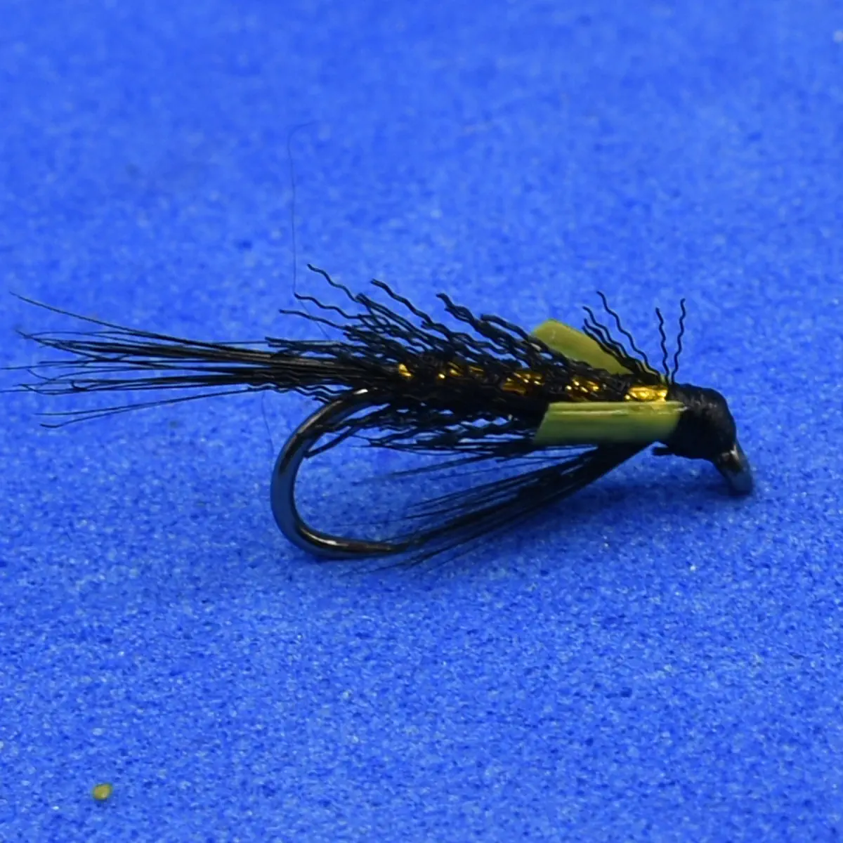 A black fishing fly lure with a curved hook, straw-colored body, and wispy tail on a blue background (angling fly)