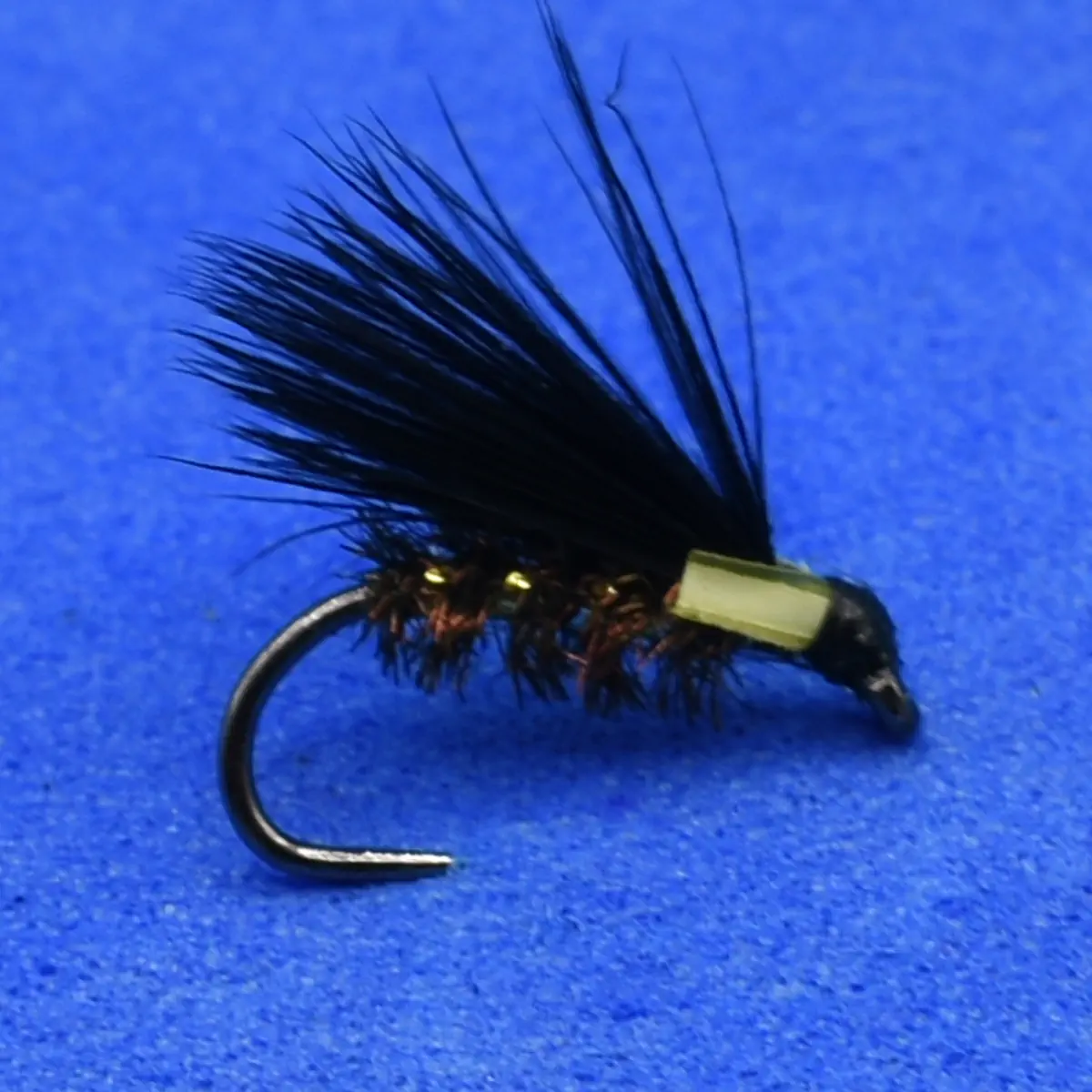 Black feathered fishing fly with a pale green body on a curved hook against a blue background.