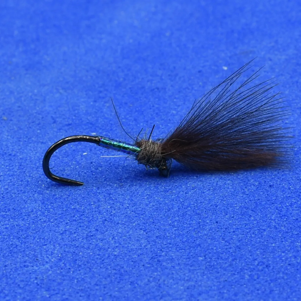 Close-up of a black fishing fly with a glossy green body and dark feather tail on a blue background.