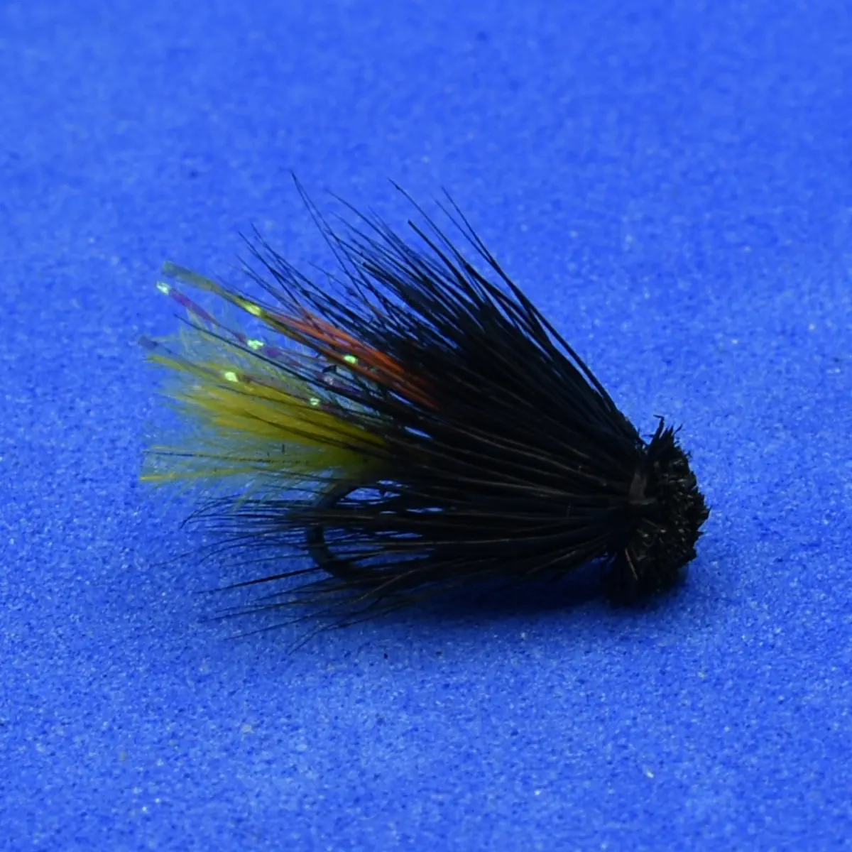 Black feathered fishing fly with yellow and orange accents on a blue background on a blue surface.
