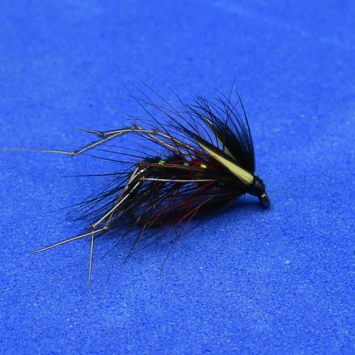 Macro shot of a tied fishing fly with black hackle, yellow tag, and iridescent bead accents against a blue background.