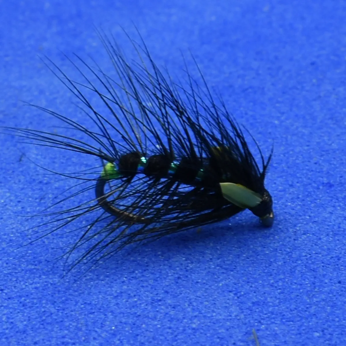 Black fishing fly with long wispy black hackle and a yellow head bead on a blue background.