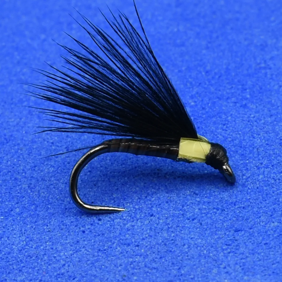 Artificial fly fishing lure: black hairy wing tied to a curved hook with a yellow head, on a blue background.
