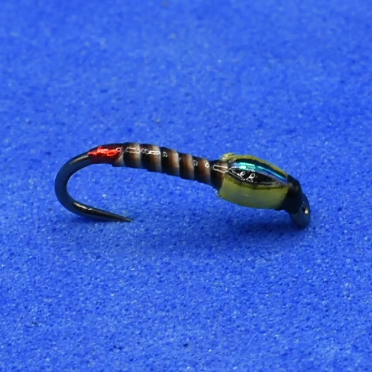 Close-up of a small fishing lure with a curved hook and segmented brown body on a blue fabric background.