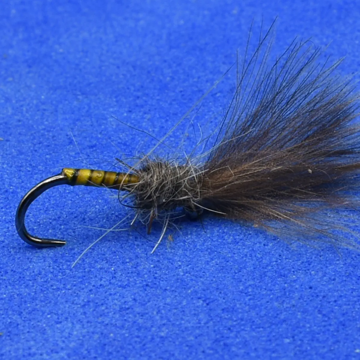 A fishing fly lure with a dark fuzzy tail and striped yellow-brown body on a curved hook, used for fly fishing.