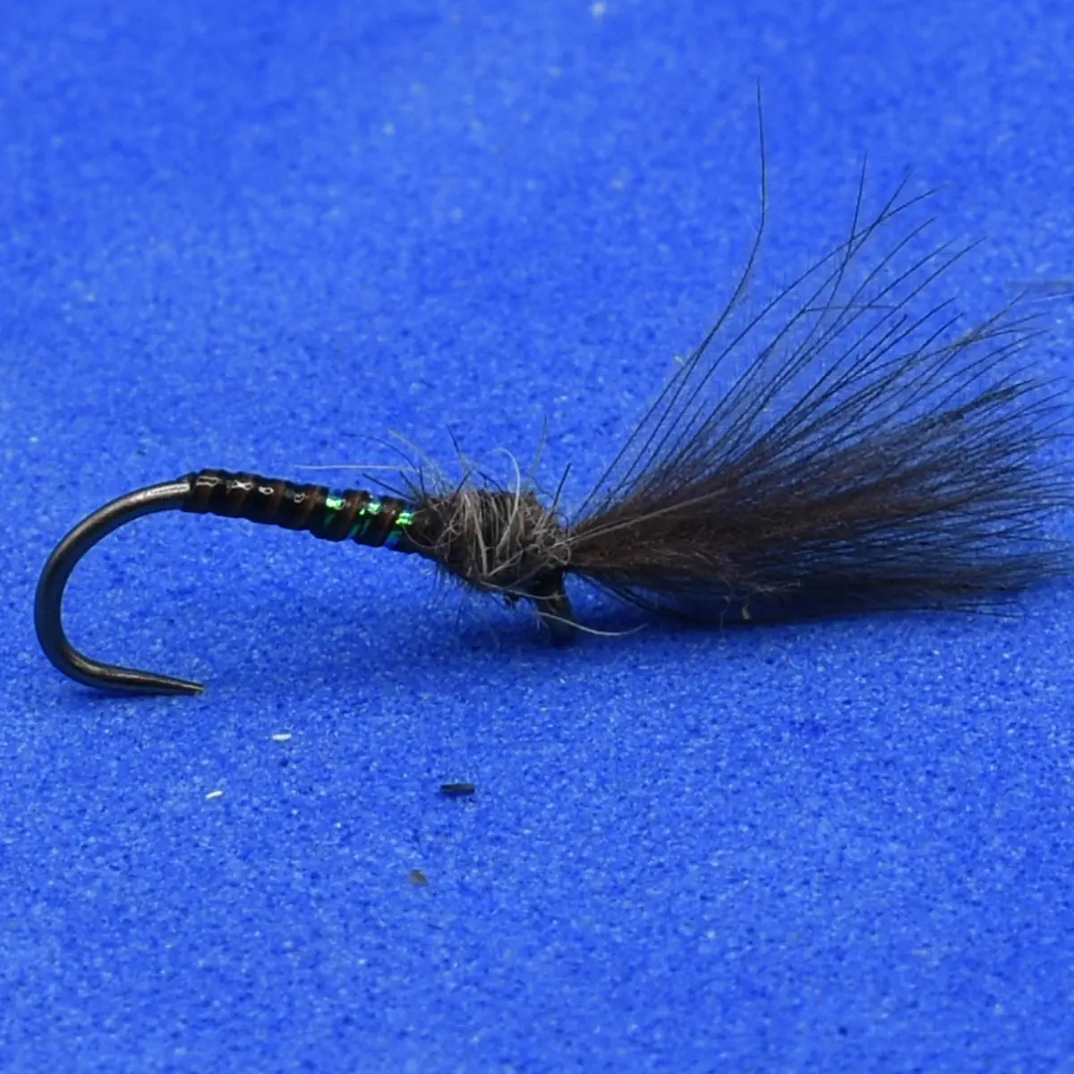 Dark fly fishing lure with a fluffy black tail and green ribbing on a curved hook, set on a blue background