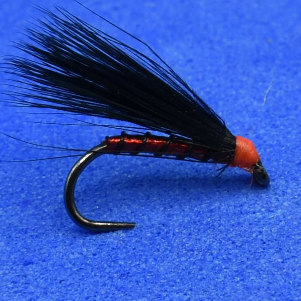 Red-bodied black streamer fishing fly with long black hackle tied to a curved hook, set on a blue background, used for fly fishing