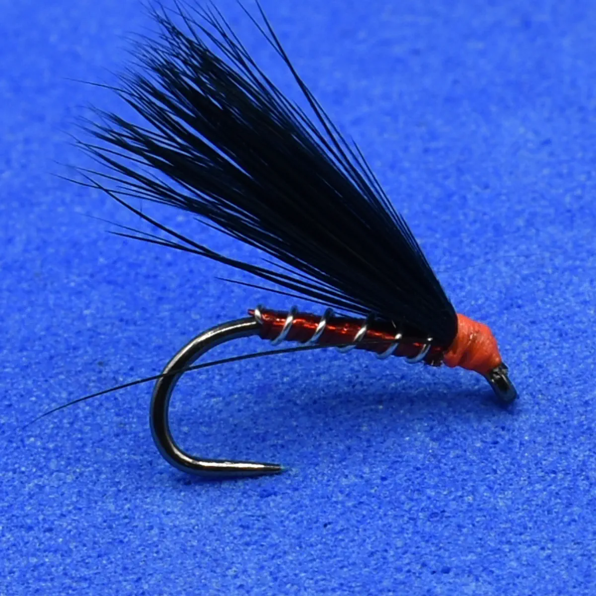 Black hackle dry fly with orange thread body mounted on a curved fishing hook against a blue background.