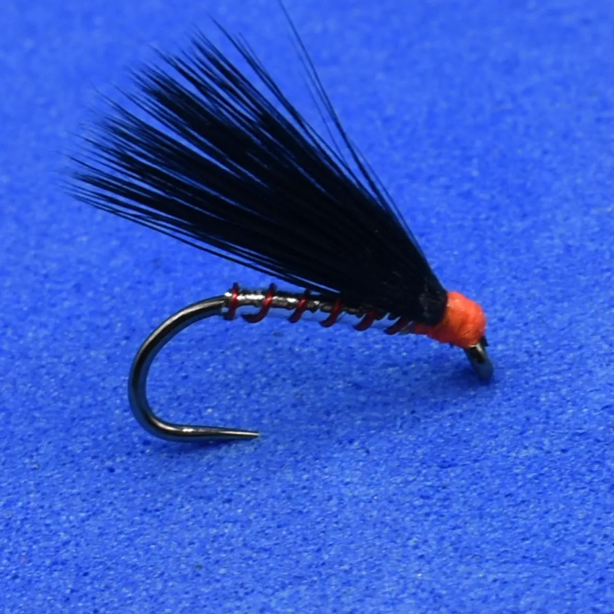 Close-up of a black feathered fishing fly tied with red thread on a curved hook, laid on a blue background.