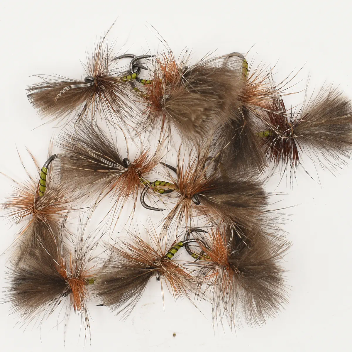 Assorted fluffy brown fishing flies with fur tails and green ribbed bodies on a white background, arranged for fly fishing.