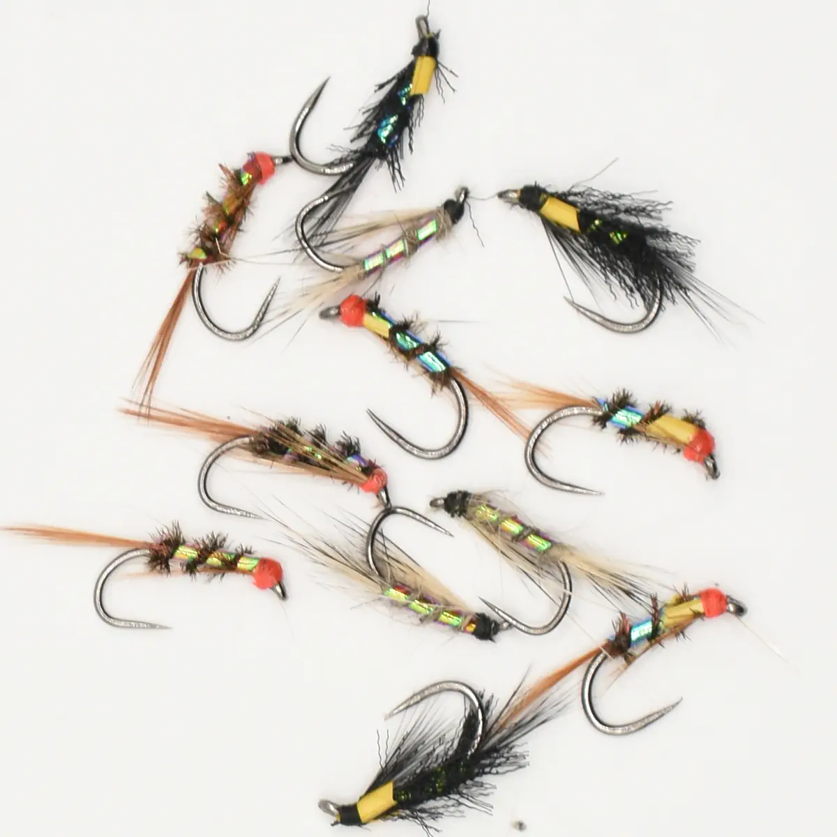 Assorted metallic fishing flies with colorful bodies scattered on a white background (fly fishing lures).
